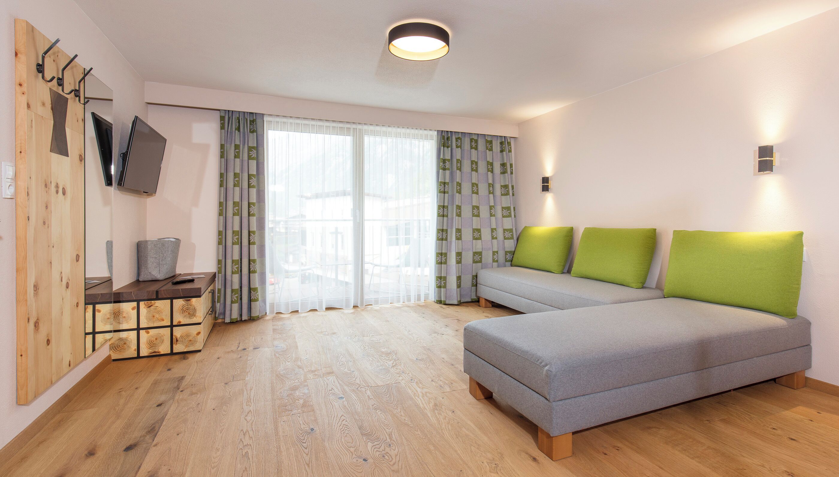 A modern bedroom with a grey sofa, green cushions, a wooden wardrobe, and a large window with curtains.