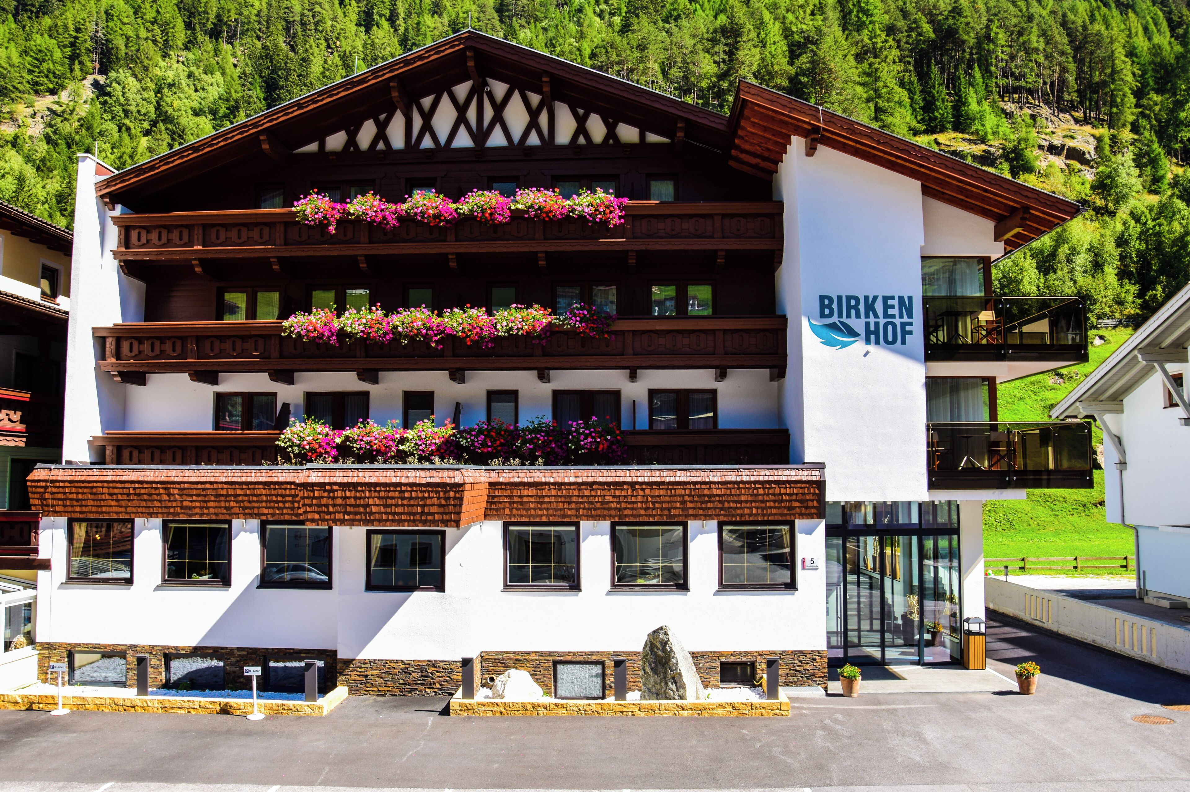 A multi-story hotel with balconies adorned with flower boxes, a sign reading 'BIRKEN HOF,' and a modern glass entrance.