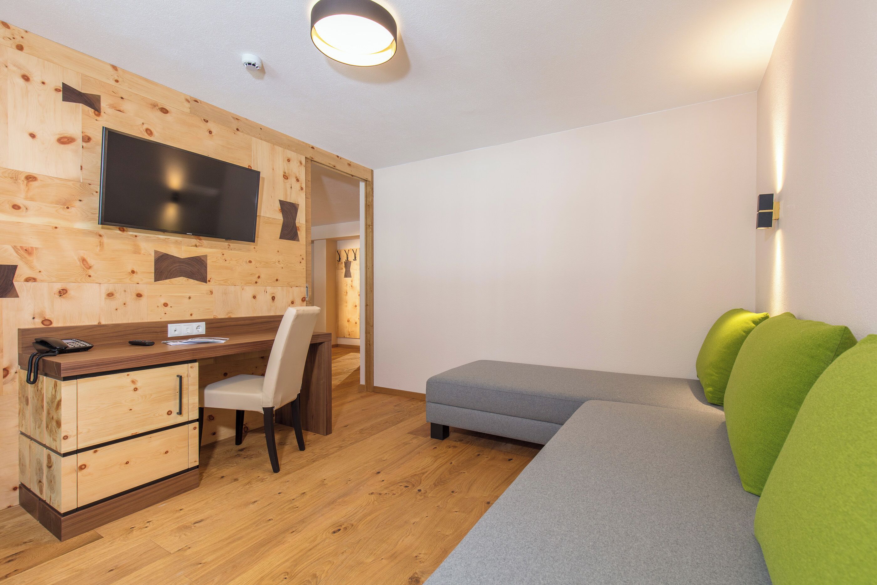 Une chambre moderne avec un mur en bois, un téléviseur à écran plat, un bureau, une chaise, un canapé gris et des coussins verts.