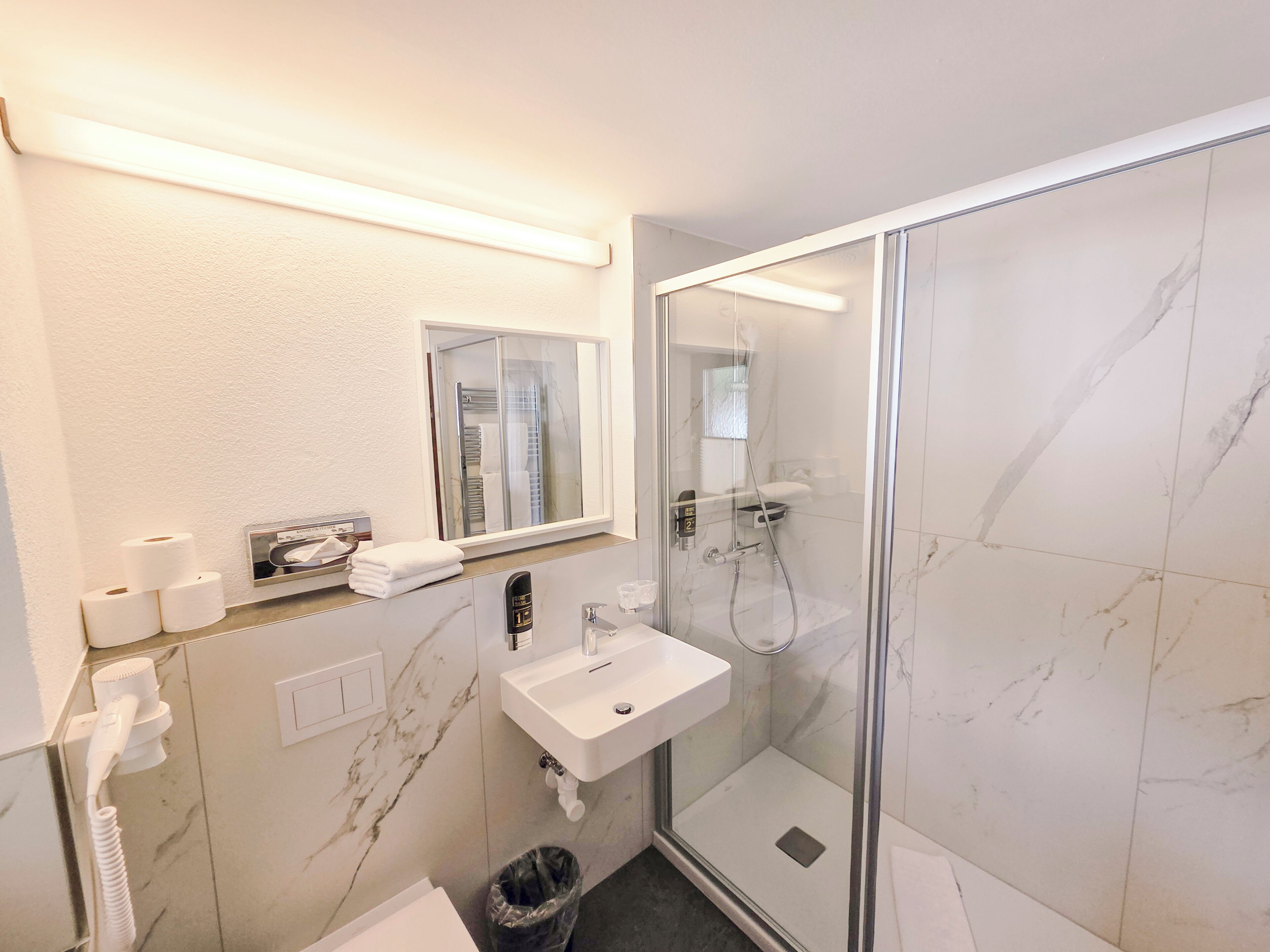 Ein modernes Badezimmer mit einer in Glas abgeschirmten Dusche, einem weißen Waschbecken und einer Wand mit Marmorplatten.