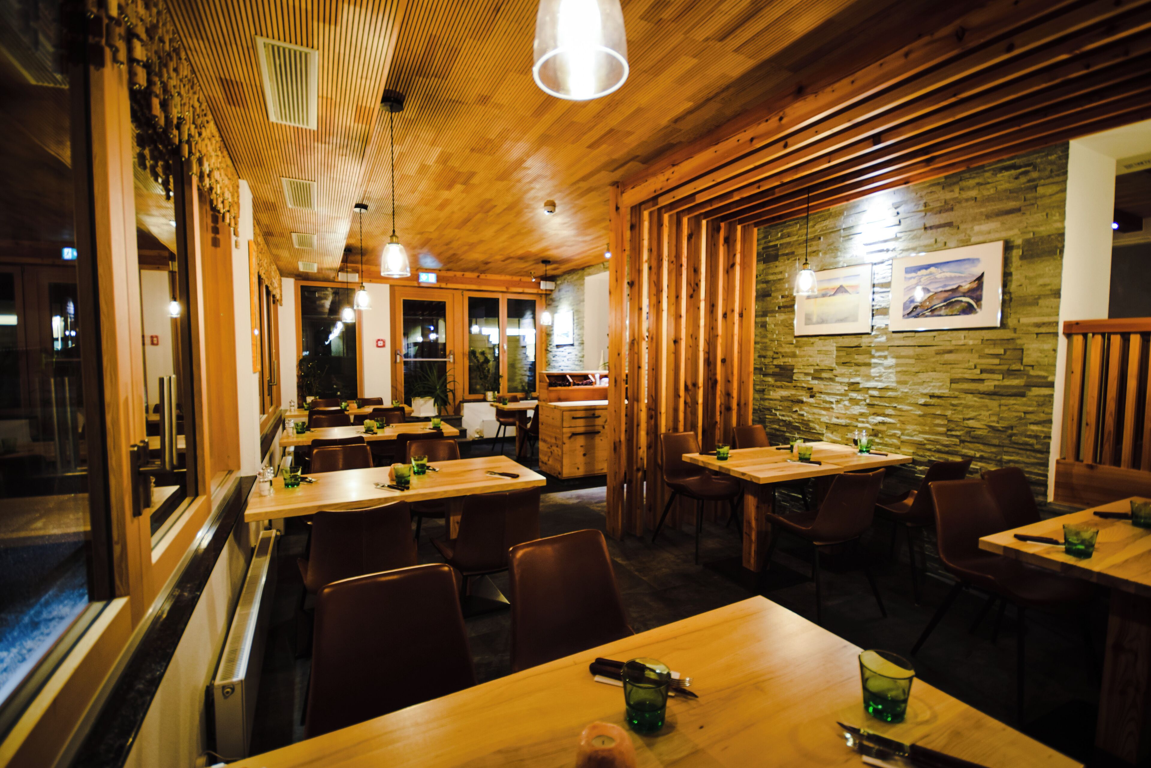 A restaurant interior with wooden tables and chairs, a stone wall, and hanging pendant lights.