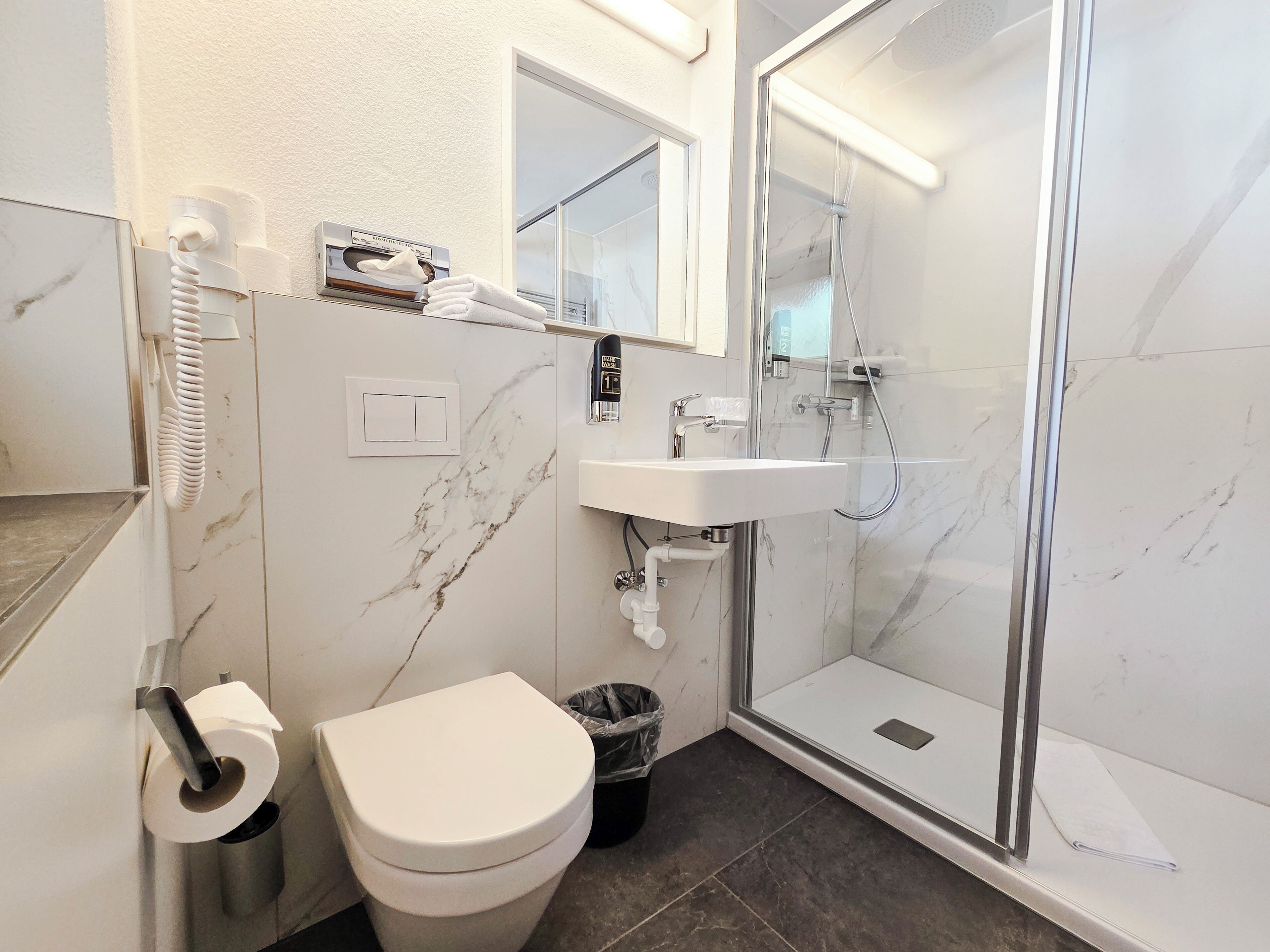A bathroom with a toilet, a sink, a shower, and a trash bin.