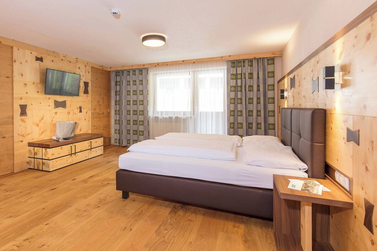 A hotel room with a wooden floor, a bed with white linens, a wooden bench with a television, and patterned curtains.