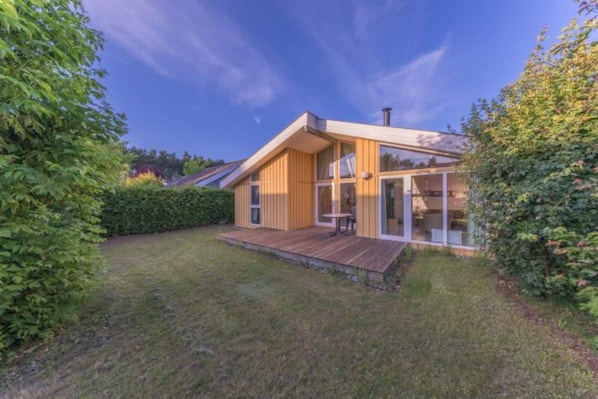 Une maison moderne dotée d’une terrasse en bois, de grandes fenêtres et d’un toit à pente.