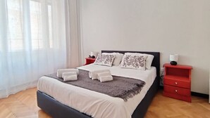 A neatly made bed with white linens and a gray throw blanket, two bedside tables with lamps, and a window with sheer curtains. - Trieste Central Holiday Home (Trieste)