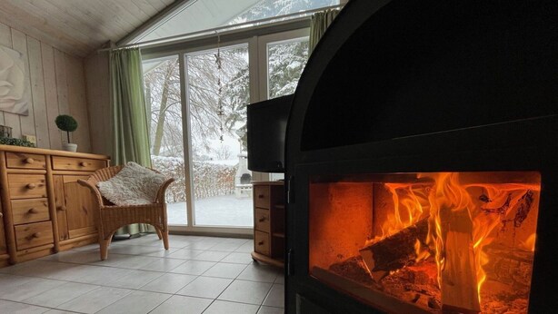 Ein gemütliches Zimmer mit Kamin, einem Korbstuhl, einer Holzkommode und Blick auf eine verschneite Landschaft durch ein großes Fenster.