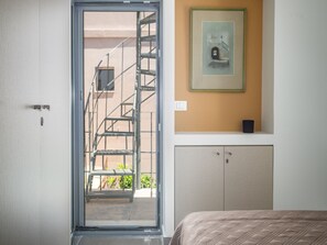 A hotel room with a sliding glass door leading to a balcony, a bed with a quilted cover, a framed picture on the wall, and a small shelf with a black object. - Elegant City Apartment - 250m From Solomos Square (Zakynthos)