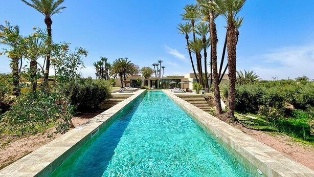 A long swimming pool with clear blue water, surrounded by palm trees and a modern building in the background. - Palais Akbal (Marrakech)