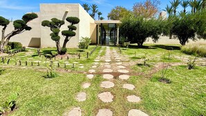 A modern building with a geometric facade, a paved pathway leading to a glass entrance, and a well-maintained garden with various shrubs and trees. - Palais Akbal (Marrakech)