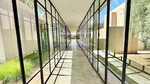 A corridor with glass walls and a tiled floor, leading to an open area with trees and a building in the background. - Palais Akbal (Marrakech)