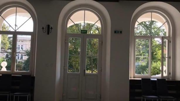 Um quarto amplo com lustre, janelas em arco e uma porta com a inscrição 'EXIT'.