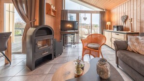 A cozy living room with a wood-burning stove, a television, wicker furniture, and a view through large windows. - Ferienhaus im Skandinavischen Stil (Rechlin)