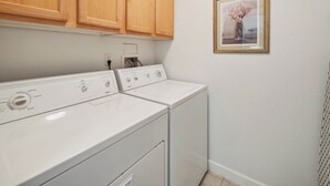 A laundry room with two white washing machines, wooden cabinets above, and a framed picture on the wall. - Havasu Hacienda Private Pool! (Lake Havasu City)