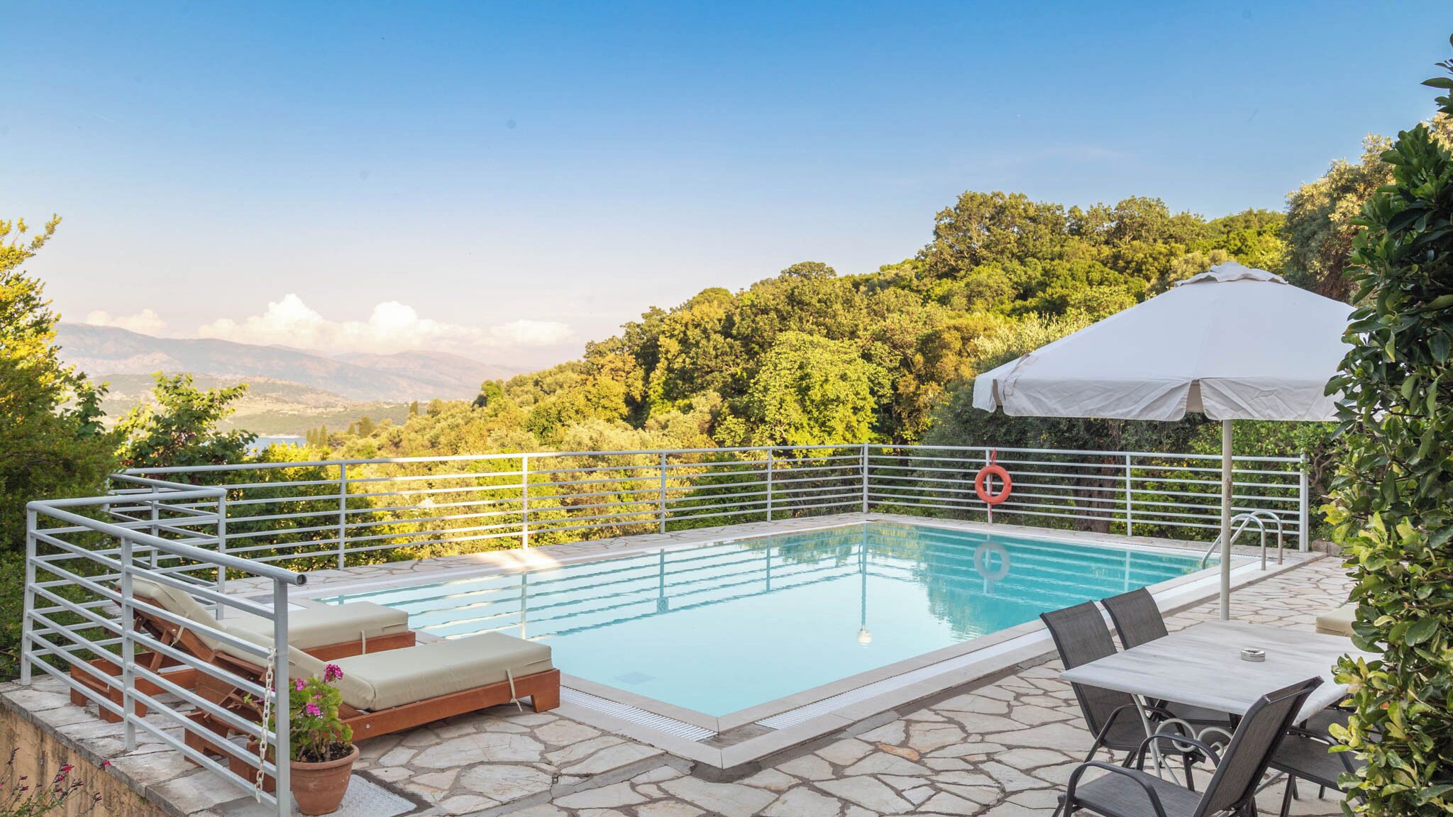 Piscina com grade de metal, espreguiçadeiras, mesa e cadeiras, e um guarda-sol branco.