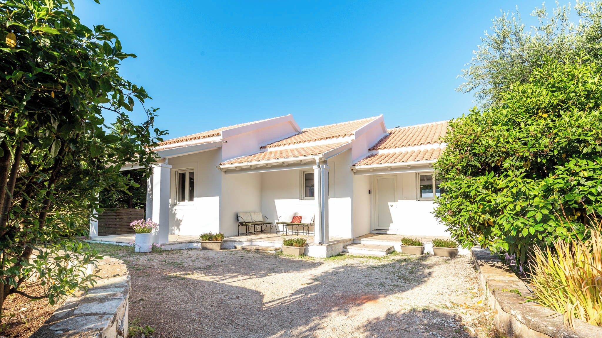 A white villa with a tiled roof, a covered porch, and a gravel driveway.