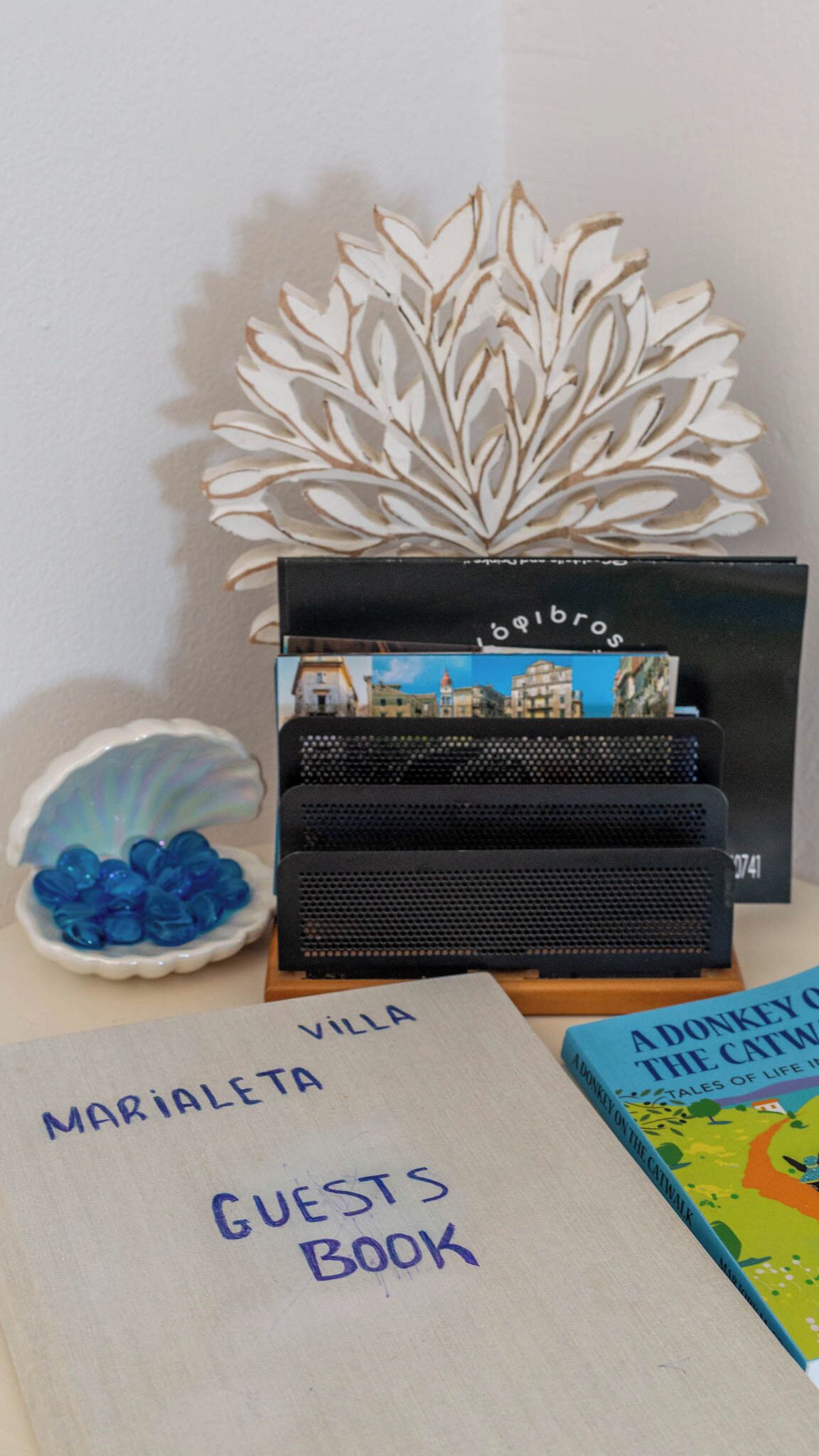 Ein Schreibtisch mit dekorativem Blattmuster, eine blaue Glasvase, ein schwarzer Lautsprecher und ein handgeschriebenes 'Guests Book' auf einer weißen Oberfläche.