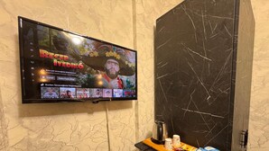 A flat-screen TV mounted on a wall displaying a video game, with a coffee table below featuring a kettle, cups, and snacks. - PENDİK SUİT OTEL (Istanbul)