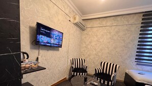 A hotel room with a marble wall, a flat-screen TV mounted above a small table, two striped armchairs, a sofa, and a window with blinds. - PENDİK SUİT OTEL (Istanbul)