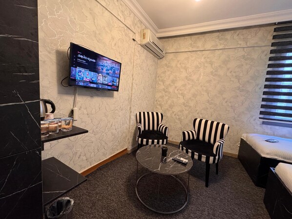 A hotel room with a marble wall, a flat-screen TV mounted above a small table, two striped armchairs, a sofa, and a window with blinds. - PENDİK SUİT OTEL (Istanbul)