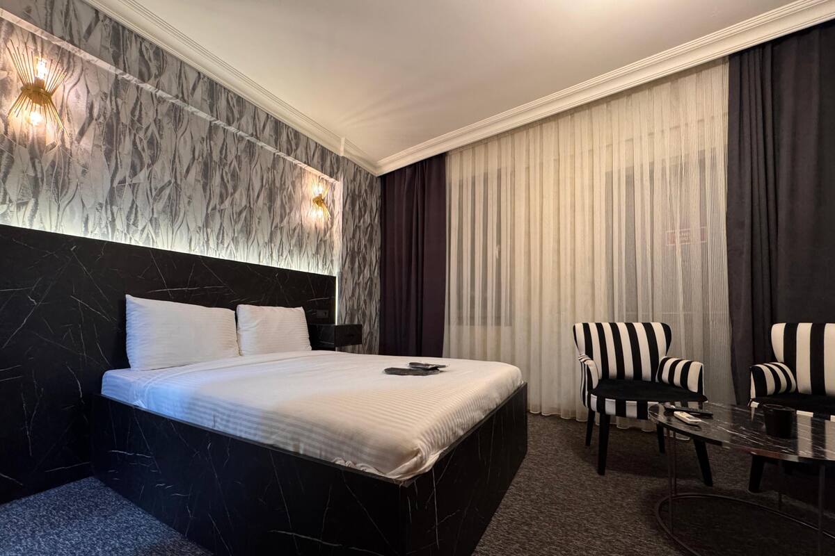 A modern hotel room with a marble headboard, a large bed, a round table, and striped armchairs.