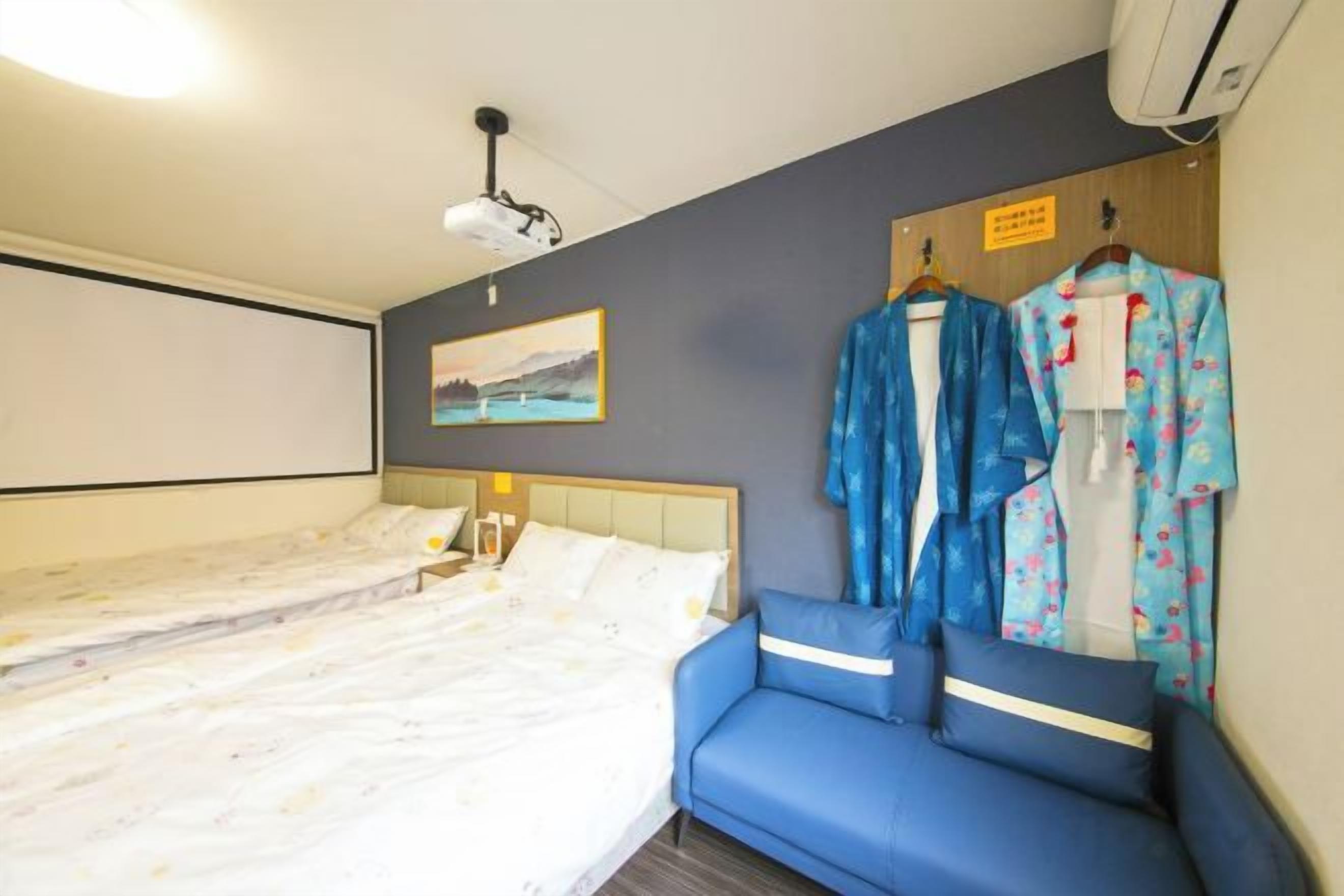 A hotel room with a bed, a blue sofa, a projector, and a wall-mounted air conditioner.