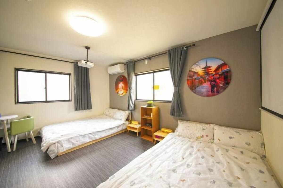 Une chambre d’hôtel avec deux lits simples, un bureau, une chaise, une petite table, une étagère et une fenêtre avec des rideaux.