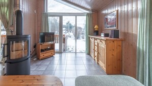 L’intérieur chaleureux d’un chalet comprend un poêle à bois, des meubles en bois, un téléviseur et une porte-fenêtre coulissante donnant sur un espace extérieur enneigé.