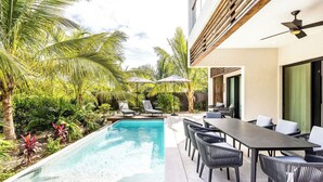 Un área junto a la piscina con una mesa larga y sillas, rodeada de exuberante vegetación y palmeras.