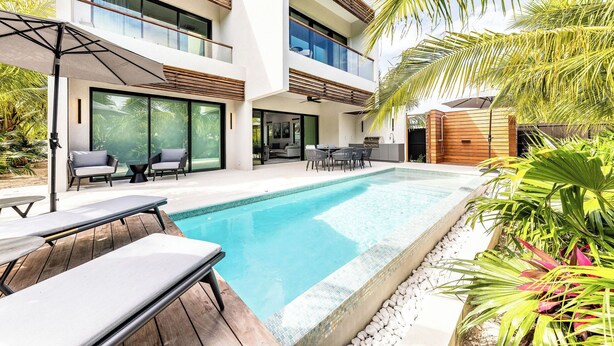 Un edificio moderno de dos pisos con piscina, mobiliario exterior y plantas tropicales.