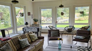 A spacious living room with a brown leather sofa, two armchairs, a coffee table, and a foosball table. Large windows offer a view of a green landscape. - World Lodge with unique, city-inspired rooms, located by the forest (Altenau)