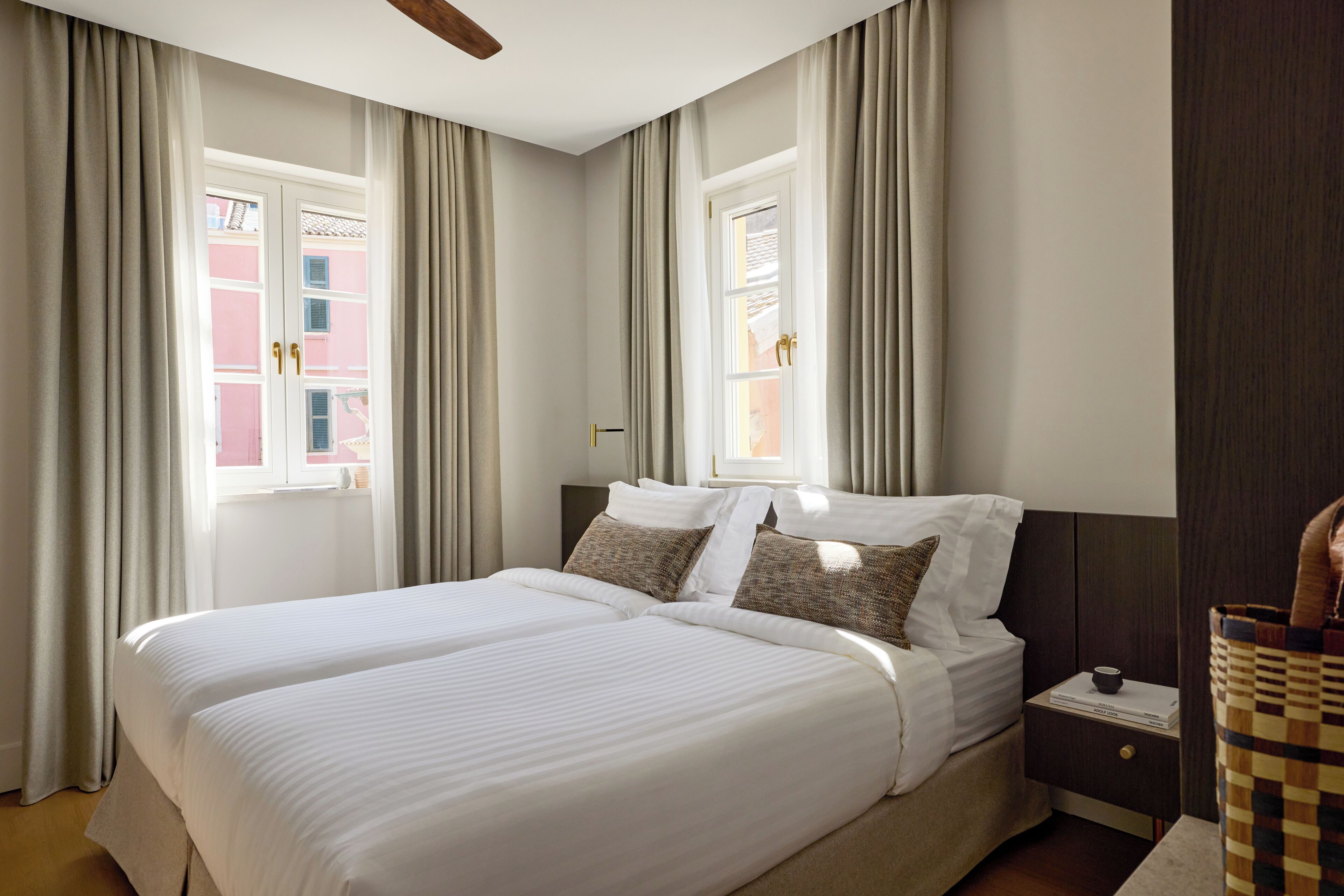 A double bed with white linens, two bedside tables with lamps, and a view of a pink building through the window.