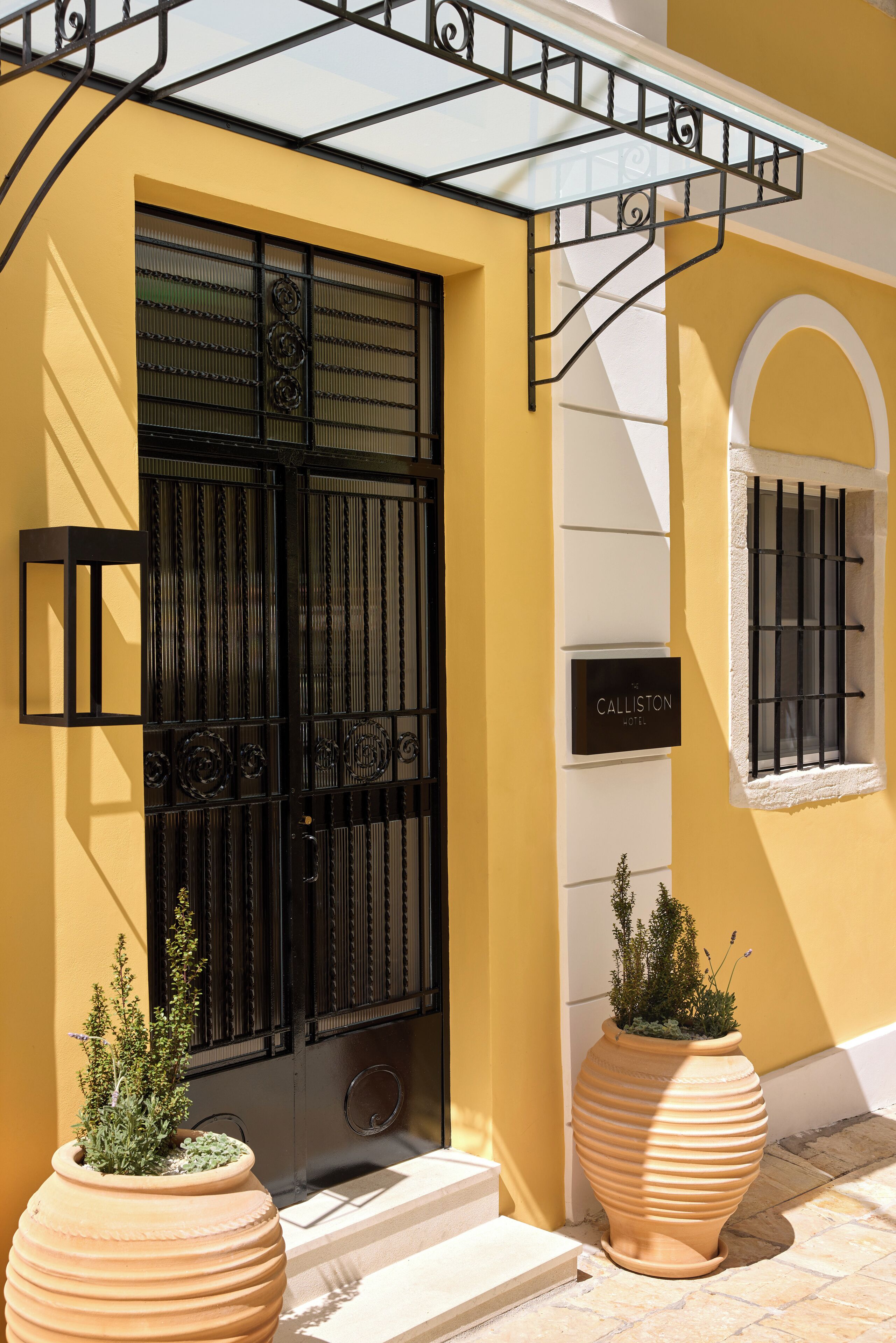 Un edificio amarillo con una reja negra, un letrero que dice "Calliston Hotel" y dos grandes macetas de terracota.