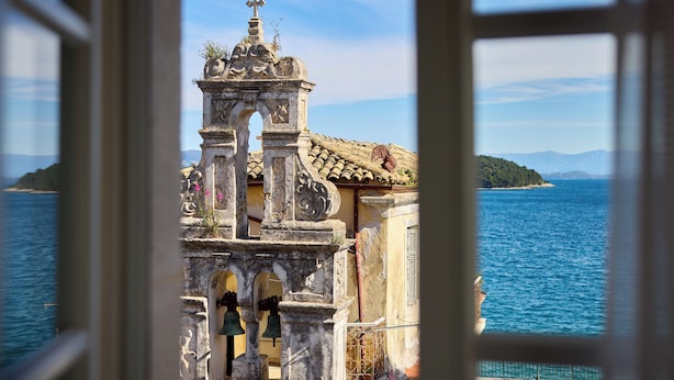 Blick durch ein Fenster auf die Küste, mit einem historischen Gebäude mit einem Glockenturm, das auf das Meer und eine entfernte Insel blickt.