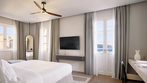 Ein modernes Hotelzimmer mit einem großen Bett, einem Deckenventilator, einem Flachbildfernseher, einem Schreibtisch mit Stuhl und Blick auf die Stadt durch große Fenster.