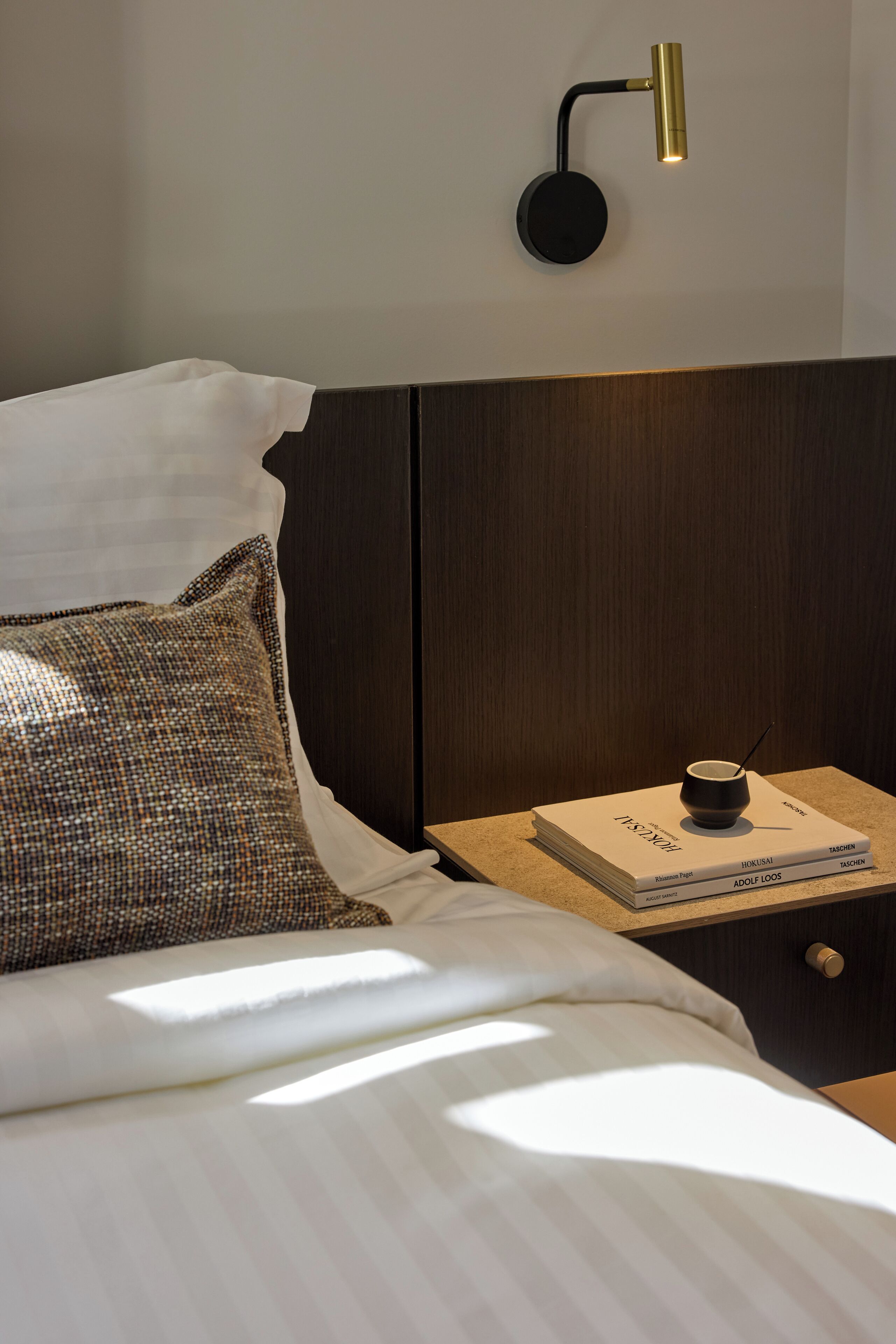 Un lit bien fait, agrémenté d’un coussin décoratif, une table de chevet avec un livre et une petite tasse, et une lampe fixée au mur.