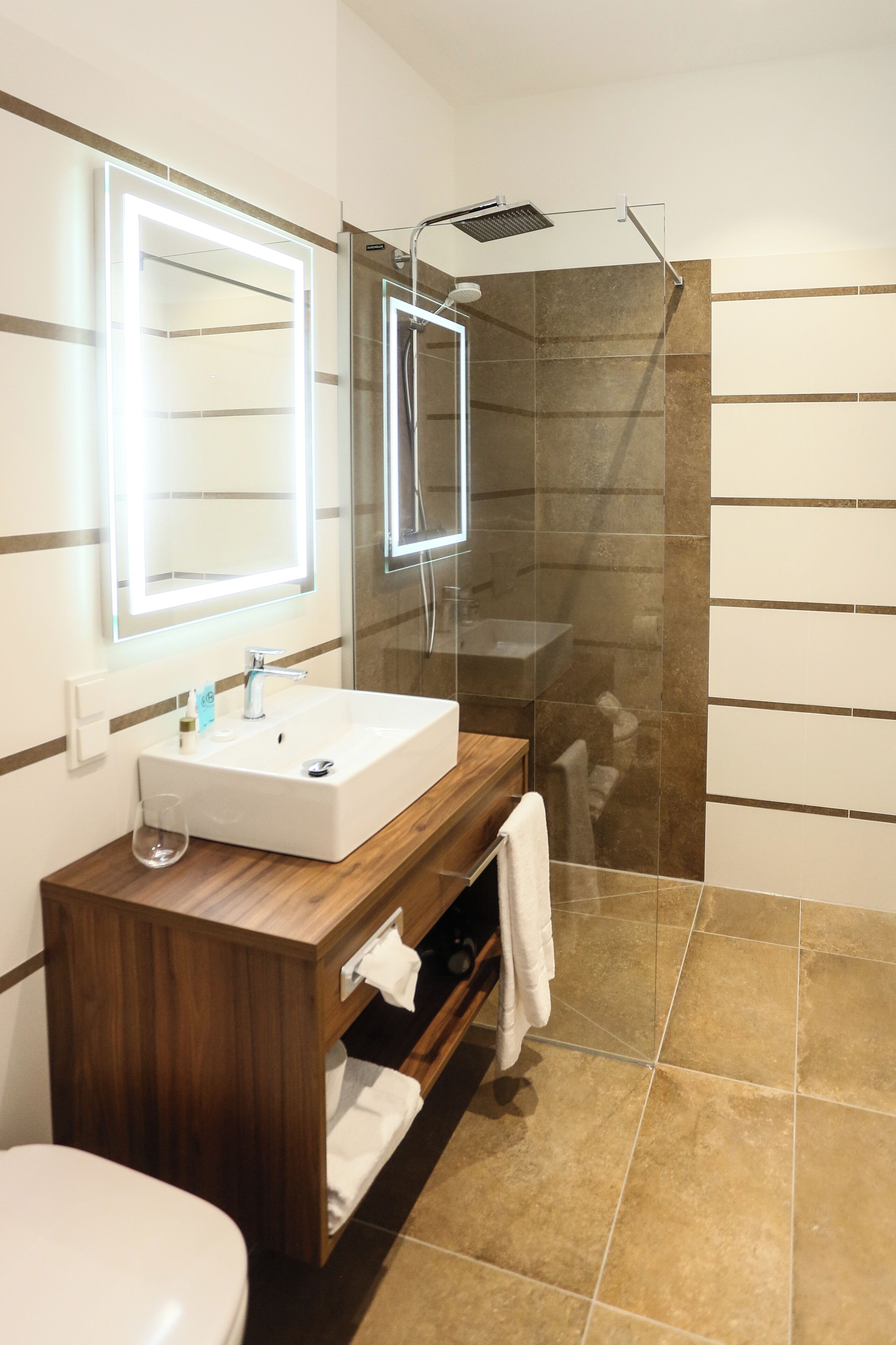 Een moderne badkamer met een glazen douchecabine, een houten wastafelmeubel met een witte wasbak en een grote spiegel met ingebouwde verlichting.