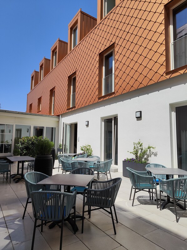 An outdoor patio area with tables and chairs, a large planter, and a building with a tiled roof and windows. - Hotel Lutz (Rain)