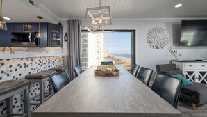 A modern dining area with a long table, bar stools, and a view of the beach. - Extraordinary 3BR Oceanfront Escape | 2 Balconies (Ocean City)
