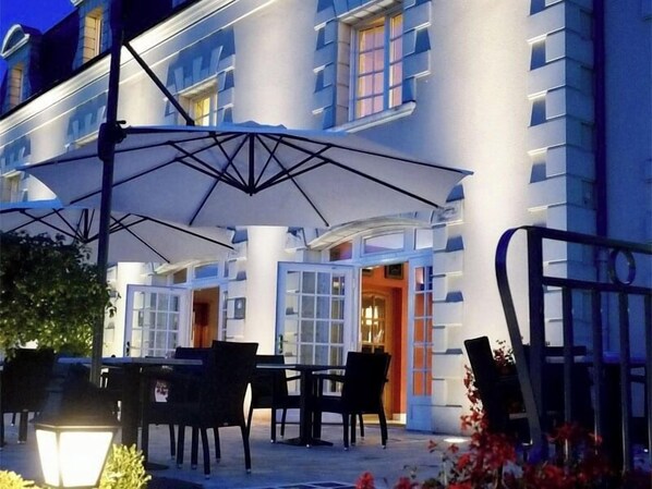 An outdoor seating area with tables and chairs, a large umbrella, and a building with multiple windows and doors.