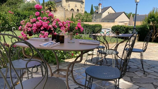 An outdoor patio with round tables and chairs, surrounded by lush greenery and pink flowers.