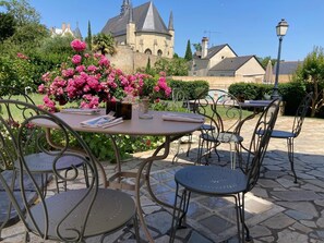 Un patio al aire libre con mesas y sillas redondas, rodeado de abundante follaje y flores rosadas.
