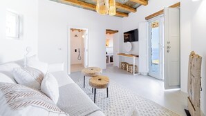 A modern, well-lit room with a wooden ceiling, a large bed with white bedding, two round stools, and a view of a beach through an open door.