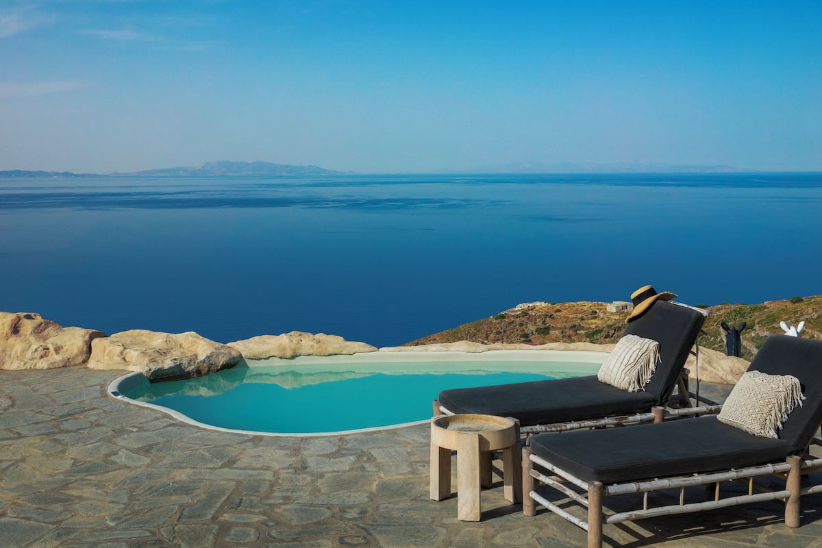 Zona de piscina con sillones de descanso y suelo de baldosas de piedra, con vistas a una extensa masa de agua y montañas lejanas.