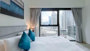 A modern hotel room with a large bed, two blue pillows, a view of tall buildings through a large window, and a cityscape. - ACTONE Tower2-Step to Burjkhalifa-1BR (Dubai)