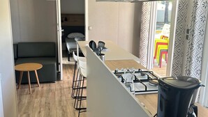A modern kitchen with a gas stove, a black trash bin, and a dining area with a table and chairs. - CAMPING 5 🌟MOBIL HOME RENTAL PREMIUM GAME AT VALRAS BEACH (Valras-Plage)