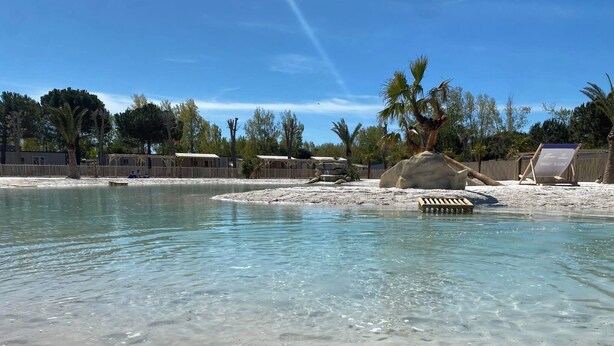 A clear pool with a sandy shore, surrounded by palm trees and a wooden fence. - CAMPING 5 🌟MOBIL HOME RENTAL PREMIUM GAME AT VALRAS BEACH (Valras-Plage)