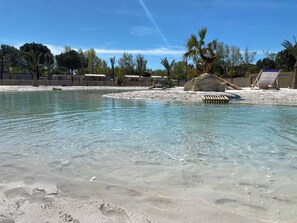 A clear pool with a sandy shore, surrounded by palm trees and a wooden fence. - CAMPING 5 🌟MOBIL HOME RENTAL PREMIUM GAME AT VALRAS BEACH (Valras-Plage)