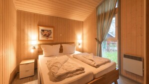 A wooden-paneled bedroom with a bed, bedside lamps, a nightstand, and a window with curtains. - Ferienhaus mit Sauna im Ferienpark Mirow (Mirow)