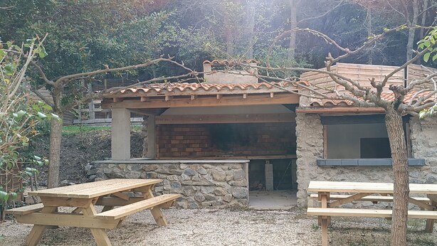 A rustic outdoor dining area with a stone wall and tiled roof, featuring two wooden picnic tables and a brick oven. - Gite d'étape Benjamin sur le sentier Cathare (3-person room) (Prugnanes)