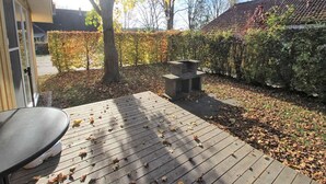 Una terraza de madera con una mesa y sillas, rodeada por un seto y árboles con hojas otoñales.